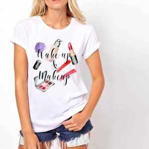 May include: White t-shirt with the text "Wake up & Makeup" in black lettering. The design features a watercolour illustration of makeup products, including lipstick, foundation, and eyeshadow.
