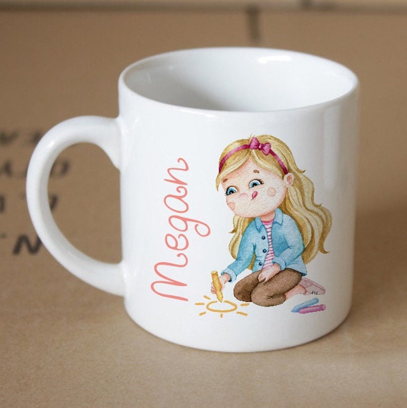 Pode incluir: Caneca de cer&acirc;mica branca com uma ilustra&ccedil;&atilde;o rosa e azul de uma menina desenhando um sol com giz. A caneca tem o nome "Megan" escrito em rosa.
