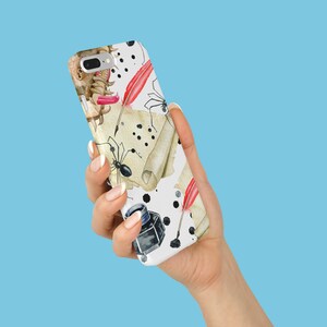 May include: A white phone case featuring a watercolor design with black ink splatters, antique scrolls, red quill pens, and black spiders. The case also includes a black inkwell and a small, brown object.