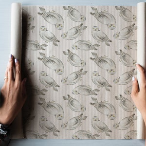 May include: A roll of wrapping paper featuring a repeating pattern of snowy owls. The owls are depicted in shades of gray and white, with yellow eyes, against a striped background. The paper is being unrolled, revealing the design.