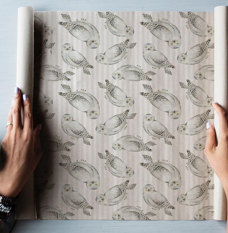 May include: A roll of wrapping paper featuring a repeating pattern of snowy owls. The owls are depicted in shades of gray and white, with yellow eyes, against a striped background. The paper is being unrolled, revealing the design.