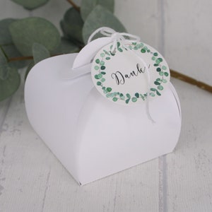 May include: A white heart-shaped gift box with a round tag. The tag has a green eucalyptus wreath design and the word "Danke" in black script. The box is on a light wood surface.