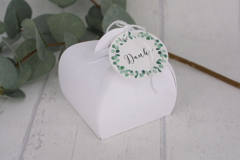 May include: A white heart-shaped gift box with a round tag. The tag has a green eucalyptus wreath design and the word "Danke" in black script. The box is on a light wood surface.