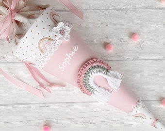 May include: A pink and white school cone with a rainbow design and a crocheted rainbow decoration. The cone is decorated with gold glitter and white polka dots. The name "Sophie" is written on the cone in white.