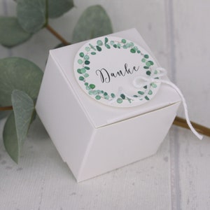 May include: A white, geometric gift box with a round tag. The tag features a green eucalyptus wreath and the word "Danke" in black script. A white string is tied to the tag and box.
