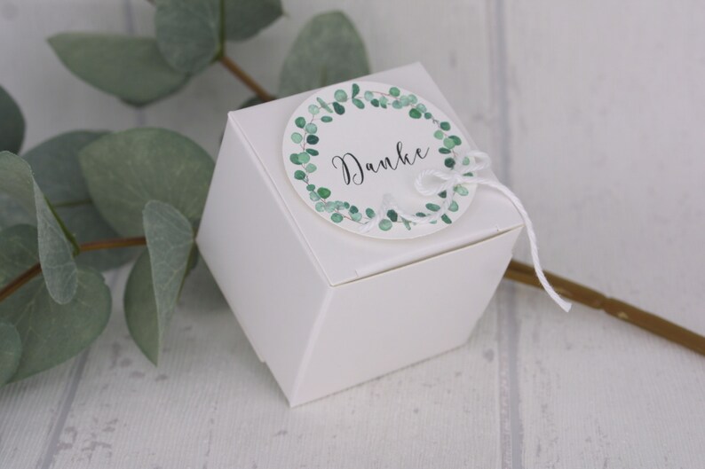 May include: A white, geometric gift box with a round tag. The tag features a green eucalyptus wreath and the word "Danke" in black script. A white string is tied to the tag and box.