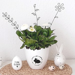Op de afbeelding: Een witte eivormige bloempot met een zwart konijnensilhouet en een bloemenkrans, met een groene plant met witte bloemen. Decoratieve paaseieren en een konijnenbeeldje zijn gerangschikt op een geweven mat.