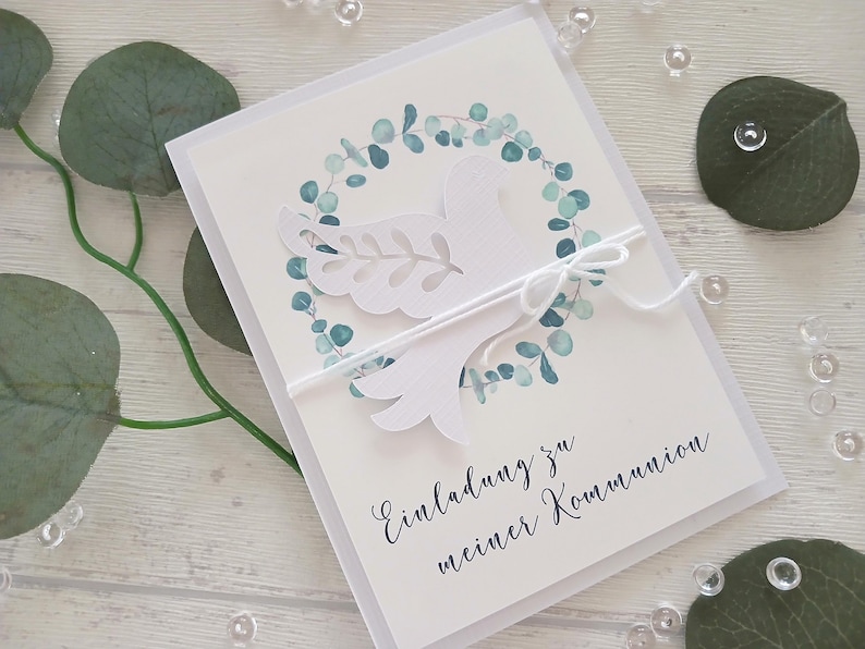 May include: White invitation card with a dove design, surrounded by a watercolor eucalyptus wreath. The card has the text "Einladung zu meiner Kommunion" in script. A white string is tied around the card.