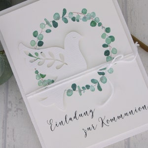 May include: White invitation card with a cut-out dove design, encircled by a watercolor eucalyptus wreath. The card is tied with white twine and features the script "Einladung zur Kommunion".