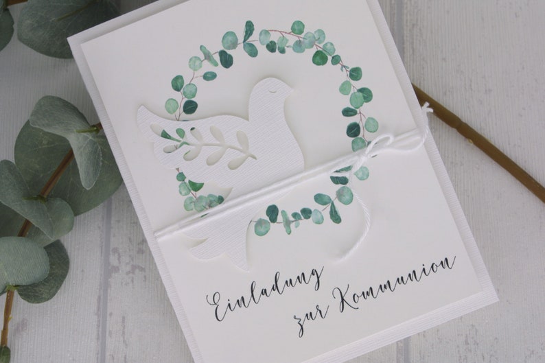 May include: White invitation card with a cut-out dove design, encircled by a watercolor eucalyptus wreath. The card is tied with white twine and features the script "Einladung zur Kommunion".