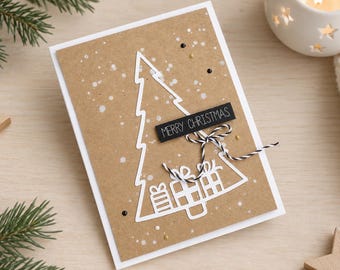 Puede incluir: Una tarjeta de felicitación rectangular con frente de papel kraft que presenta un contorno de árbol de Navidad troquelado en blanco con cajas de regalo. Una etiqueta negra dice MERRY CHRISTMAS, decorada con un lazo de cuerda bicolor.