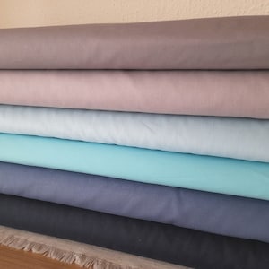 Może przedstawiać: Stosik sześciu rolek tkaniny w różnych odcieniach niebieskiego i szarego. Rolki są ułożone od najciemniejszego do najjaśniejszego, z najciemniejszą rolką na dole i najjaśniejszą rolką na górze.