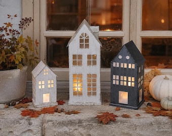 Puede incluir: Tres farolillos con forma de casa con ventanas y puertas recortadas. Dos farolillos tienen un acabado en pintura blanca desgastada, mientras que el tercero es de color negro mate. Cada estructura cuenta con iluminación interna.