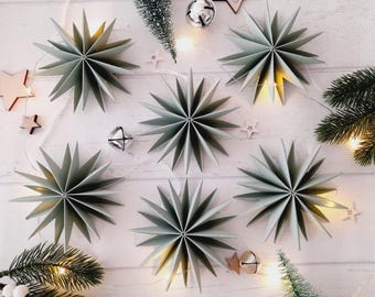 May include: A top-down view of six sage green paper star decorations, alongside small wooden stars, silver bells, and evergreen sprigs on a white wooden surface. Fairy lights provide a warm glow to the festive scene.