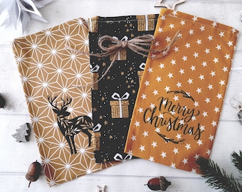 May include: Three fabric gift bags with festive patterns. One bag features a geometric design with a deer silhouette, another has a black background with gold gift boxes, and the third has a star pattern with "Merry Christmas" text.