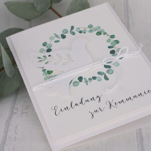 May include: A white invitation card with a cut-out dove design, encircled by a watercolor eucalyptus wreath. The card is tied with white twine and features the text "Einladung zur Kommunion" in elegant script. Green eucalyptus leaves are visible.