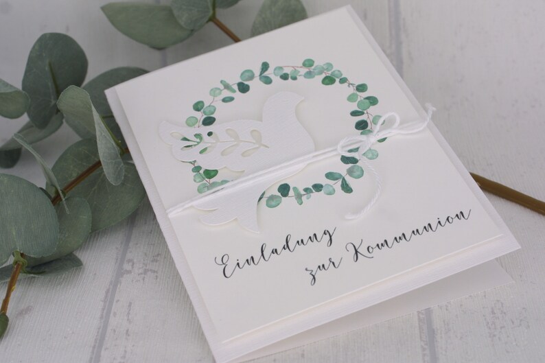 May include: A white invitation card with a cut-out dove design, encircled by a watercolor eucalyptus wreath. The card is tied with white twine and features the text "Einladung zur Kommunion" in elegant script. Green eucalyptus leaves are visible.