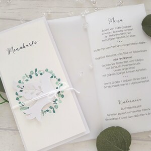May include: White wedding menu card with a dove and eucalyptus wreath design. The menu card is tied with a white string. The menu is printed in black script. The card is open, revealing the menu details.