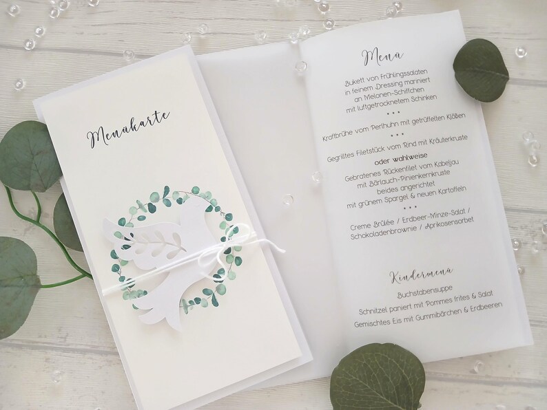 May include: White wedding menu card with a dove and eucalyptus wreath design. The menu card is tied with a white string. The menu is printed in black script. The card is open, revealing the menu details.