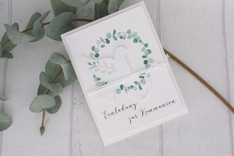 May include: White invitation card with a dove and eucalyptus wreath design. The card features the text "Einladung zur Kommunion" and is tied with white twine. Green eucalyptus leaves are visible.