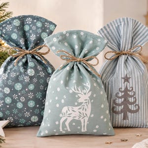 Könnte beinhalten: Drei Stoffgeschenktüten, die mit Bindfaden verschlossen sind. Die linke Tüte ist dunkelgrau mit hellblauen Schneeflocken. Die mittlere Tüte ist hellgrün mit einem weißen Hirsch und Schneepunkten. Die rechte Tüte hat graue Streifen und einen Baum.