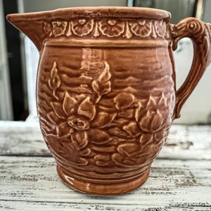 May include: Brown ceramic pitcher with a detailed floral relief design. The pitcher has a spout, a decorative handle, and a wide base. The floral pattern is repeated around the body of the pitcher. The pitcher is likely vintage or antique.