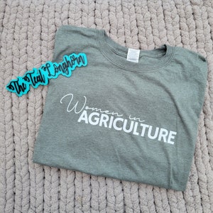 May include: A green t-shirt with the text "Women in Agriculture" printed in white. The t-shirt is on a fuzzy, light brown surface. A teal blue sticker with the text "The Teal Longhorn" is in the upper left corner of the image.