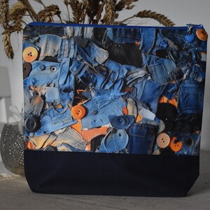 Könnte beinhalten: Eine quadratische Patchwork-Tasche aus Jeansstoff mit marineblauem Boden und blauem Reißverschluss. Die Tasche ist mit orangefarbenen Knöpfen und einem herzförmigen blauen Anhänger verziert. Das Patchwork-Design besteht aus verschiedenen Blautönen.