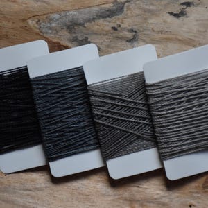 Könnte beinhalten: Vier Garnspulen in verschiedenen Grautönen, von dunkler Holzkohle bis hellgrau, werden auf weißen Karten präsentiert. Das Garn ist eng gewickelt, und die Karten sind auf einer Holzoberfläche angeordnet, was eine neutrale und strukturierte Ästhetik erzeugt.