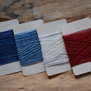 Könnte beinhalten: Vier Garnspulen in Blau-, Weiß- und Rottönen. Jede Spule ist um eine weiße rechteckige Karte gewickelt. Die Fäden sind auf einer Holzoberfläche angeordnet und zeigen die Farben und Texturen des Garns.