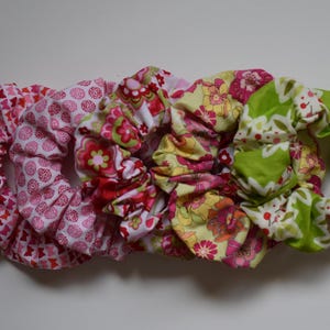 Könnte beinhalten: Eine Sammlung von fünf Stoff-Haargummis in verschiedenen floralen und geometrischen Mustern. Die Haargummis weisen Farbtöne von Rosa, Rot, Gelb und Grün auf. Diese Haaraccessoires sind dazu bestimmt, das Haar an Ort und Stelle zu halten.