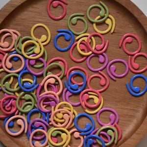 Könnte beinhalten: Ein Holztablett gefüllt mit farbenfrohen spiralförmigen Maschenmarkierern in Rosa, Gelb, Blau, Grün und Lila. Diese Strickaccessoires sind auf einer braunen Holzoberfläche angeordnet.