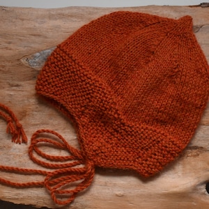 Könnte beinhalten: Eine gestrickte Babyhaube in einer verbrannten Orangetönung mit einer langen Schleife. Die Haube ist aus einem weichen, strukturierten Garn gefertigt und hat ein schlichtes, klassisches Design.