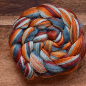 Könnte beinhalten: Ein runder Zopf aus farbenfrohem Wollvlies in Orange-, Türkis-, Burgunder- und Weißtönen. Die Faser ist fest gesponnen und kreisförmig angeordnet, ideal für Spinn- oder Filzprojekte.