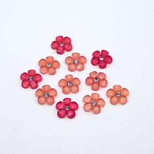 May include: A collection of ten small, pink and orange flower-shaped embellishments with a single rhinestone in the center of each flower.