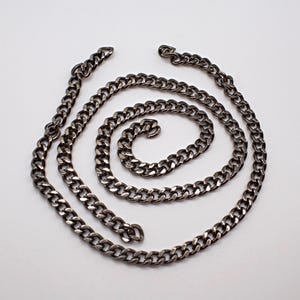 May include: A silver-coloured curb chain necklace, arranged in a spiral on a white background. The chain links are closely connected, creating a continuous, textured surface. The necklace is a simple, yet bold accessory.