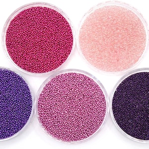 May include: Five small containers filled with colourful beads. The beads are in shades of pink, purple, and dark purple.