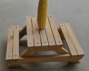 May include: A miniature wooden picnic table with attached benches. A single ear of yellow corn stands upright on the table's surface. The table is constructed from light-colored wood and secured with screws.