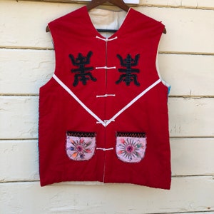 May include: A red quilted vest with white piping and two black embroidered characters on the front. The vest has two pockets with embroidered floral designs.