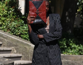 May include: A black and rust-colored leather clutch bag with a triangular flap closure, held in front of a black pinstriped blazer with white vertical lines, featuring a double-breasted button front and long sleeves.