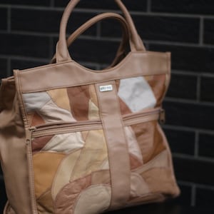 May include: A tan leather tote bag with a patchwork design in various shades of brown and beige. The bag has two handles, a zip pocket, and a small metal logo. The bag is a medium size.