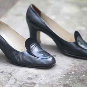 Könnte beinhalten: Ein Paar schwarze Lederschuhe mit Absatz, spitzer Zehenpartie und einem dekorativen Überlapp am Oberteil. Die Schuhe haben einen niedrigen Blockabsatz.