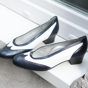 Può includere: Un paio di scarpe bicolore con tacco basso. Le scarpe presentano una tomaia bianca con dettagli blu navy e un design traforato decorativo sulla punta. I tacchi sono blu navy scuro. Le scarpe sono appoggiate su una superficie bianca.