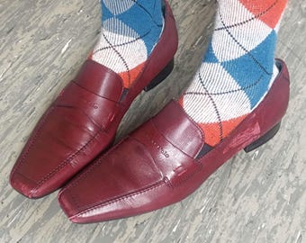 May include: A pair of dark red leather loafers with a square toe and a strap across the vamp featuring the Marc O'Polo logo. The shoes are worn with white, orange, and blue argyle patterned socks that reach above the ankle.