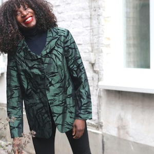 Pode incluir: Um blazer de manga comprida com uma base verde-escura e um padrão preto abstrato e orgânico. O casaco tem um colarinho largo, fecho frontal com botões e um corte estruturado, usado sobre uma blusa preta de gola alta e calças pretas.