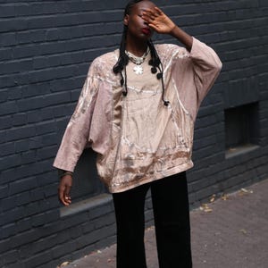Könnte beinhalten: Ein beiges, locker sitzendes Langarmshirt mit strukturierter, schimmernder Oberfläche und weit fallender Silhouette. Das Kleidungsstück hat einen Rundhalsausschnitt und wird mit einer schlichten schwarzen Hose kombiniert. Eine silberne Kette.