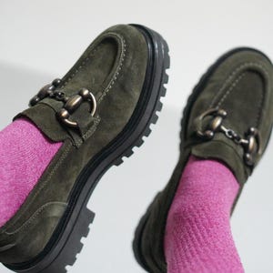 May include: A pair of olive green suede loafers featuring a prominent dark metal horsebit detail across the vamp, thick black lug soles with deep treads, and visible stitching along the toe box, worn with bright pink ribbed knit socks.