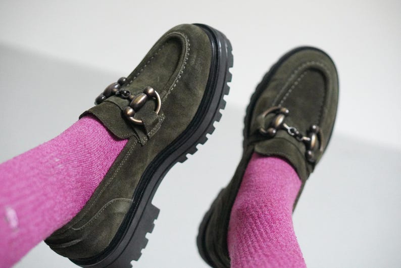 May include: A pair of olive green suede loafers featuring a prominent dark metal horsebit detail across the vamp, thick black lug soles with deep treads, and visible stitching along the toe box, worn with bright pink ribbed knit socks.