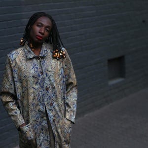 Op de afbeelding: Een vrouw met lang, donker haar draagt een bruin en blauw gepatroneerd jack met houten kralen. Ze staat voor een grijze bakstenen muur.