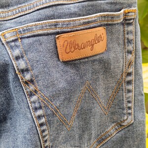 Pode incluir: Close-up de um bolso de cal&ccedil;as de ganga azul com uma etiqueta de couro castanho "Wrangler". O bolso apresenta um design de costura em "W" cl&aacute;ssico e &eacute; acentuado com costuras laranja. As cal&ccedil;as de ganga est&atilde;o desbotadas e gastas.